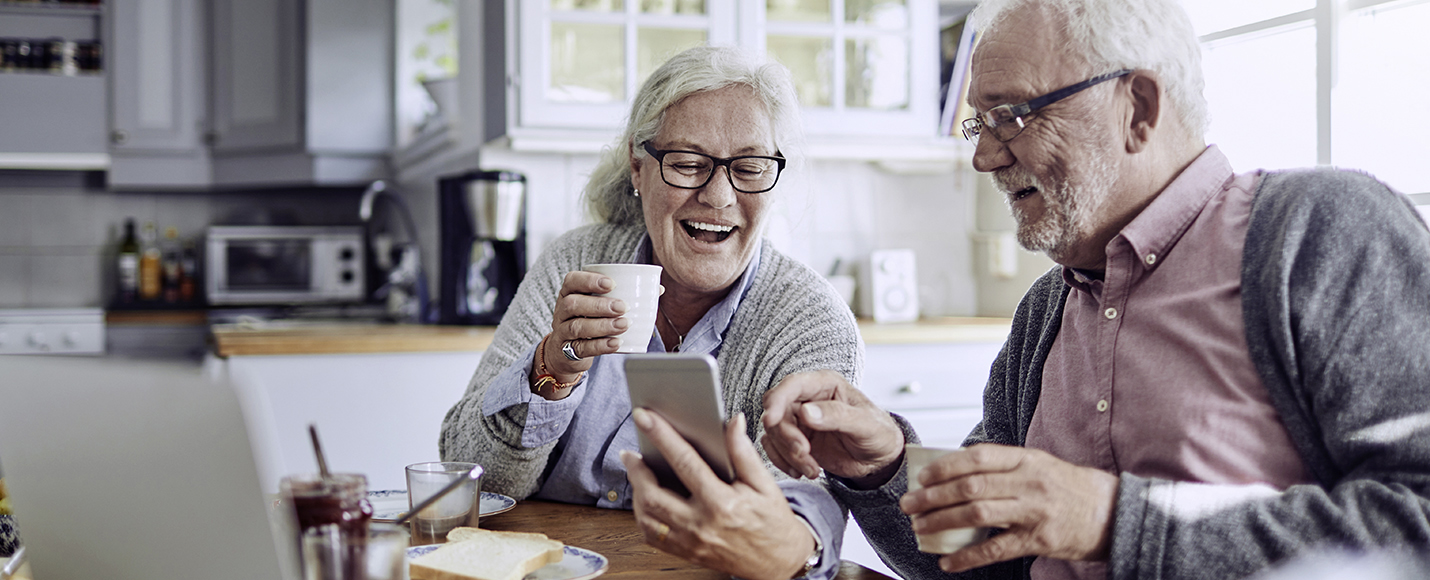 Elderly couple sitting in kitchen having a coffee looking at smartphone