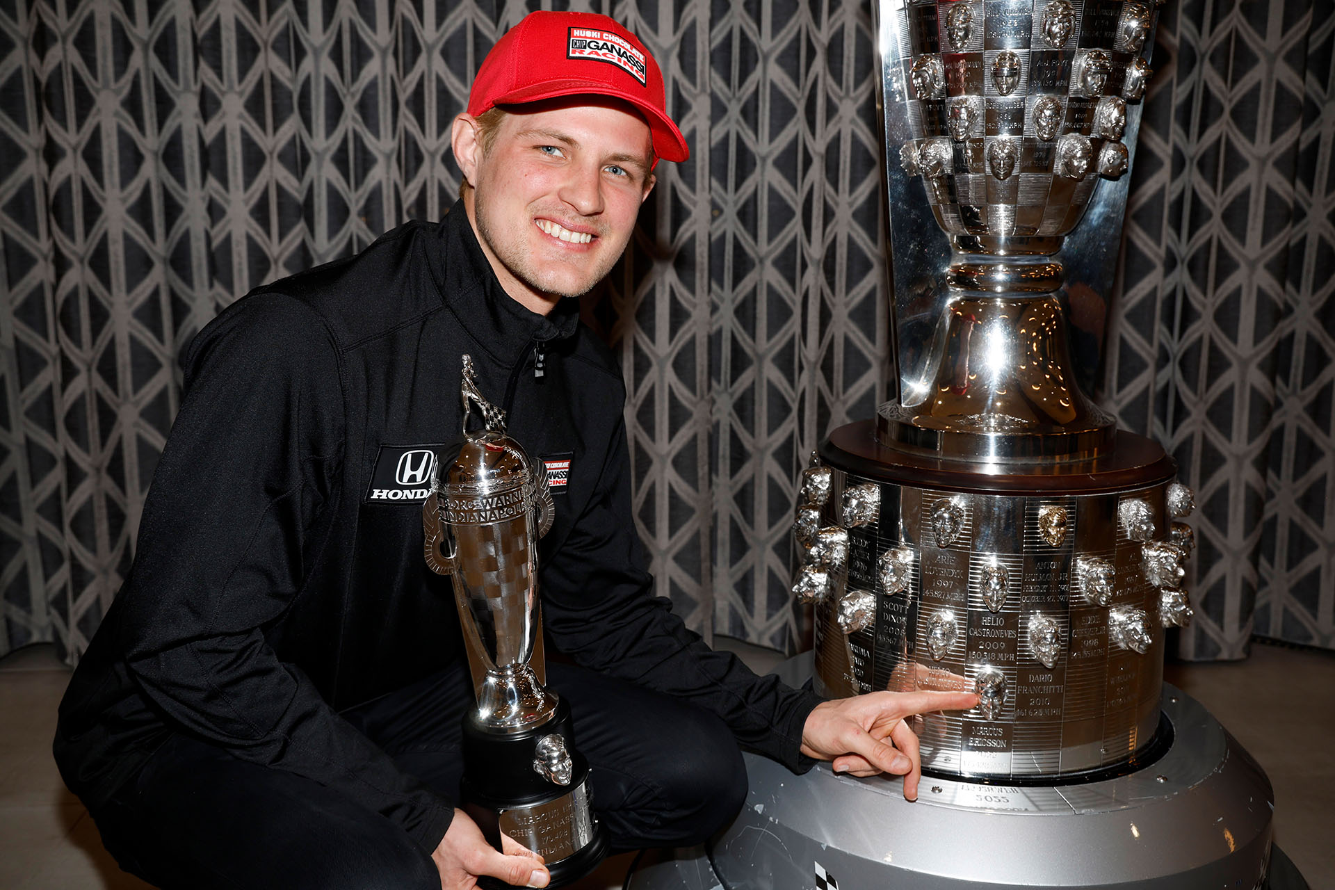 Smiling man holding trophy points at larger trophy