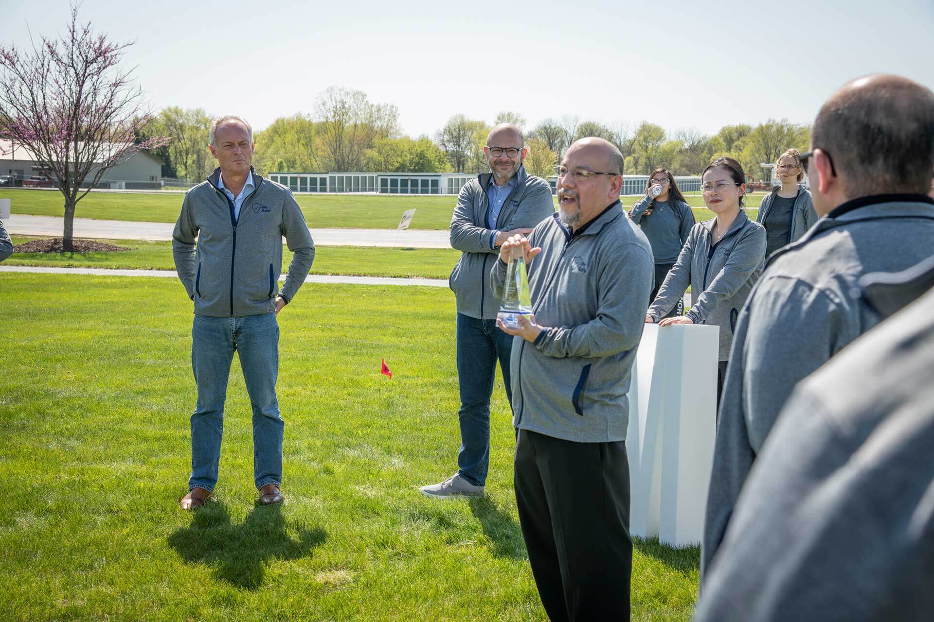 Man speaks while holding trophy in field surrounded by other people