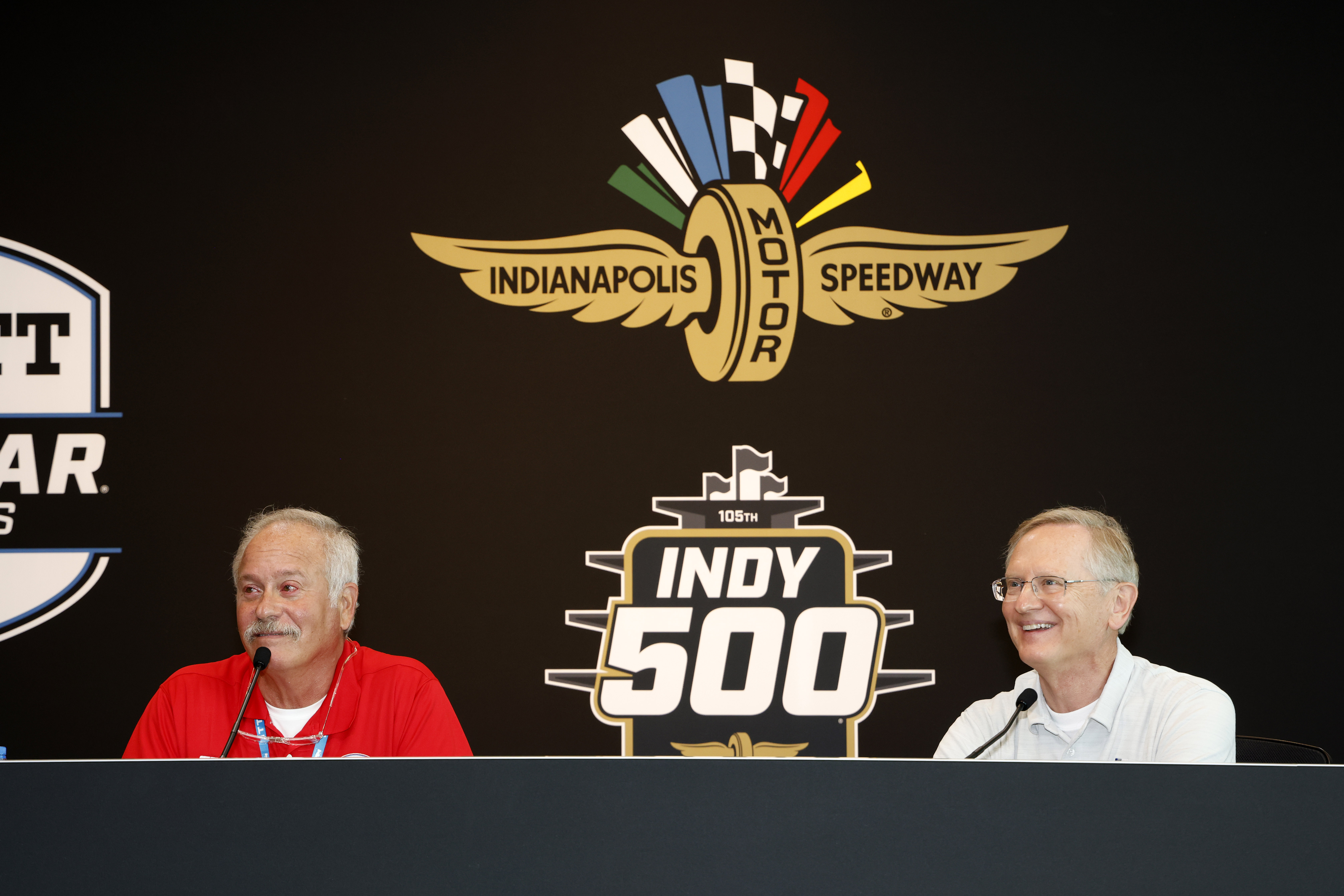 Two men sit behind table with microphones against black logo backdrop